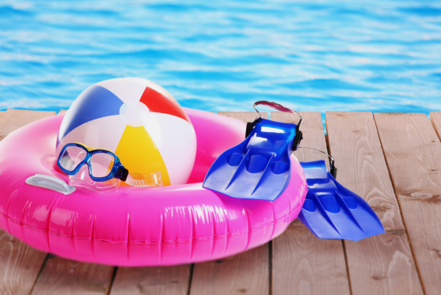 Schwimmreifen, Schwimmflossen, Taucherbrille und Wasserball liegen auf einem Steg vor einem Pool.