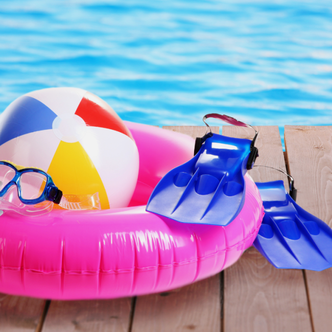 Schwimmreifen, Schwimmflossen, Taucherbrille und Wasserball liegen auf einem Steg vor einem Pool.